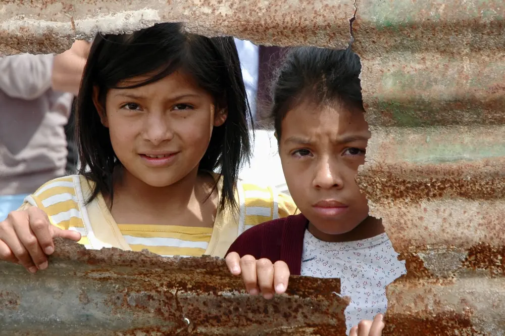 Two children peering out through a gap in a corrugated metal wall