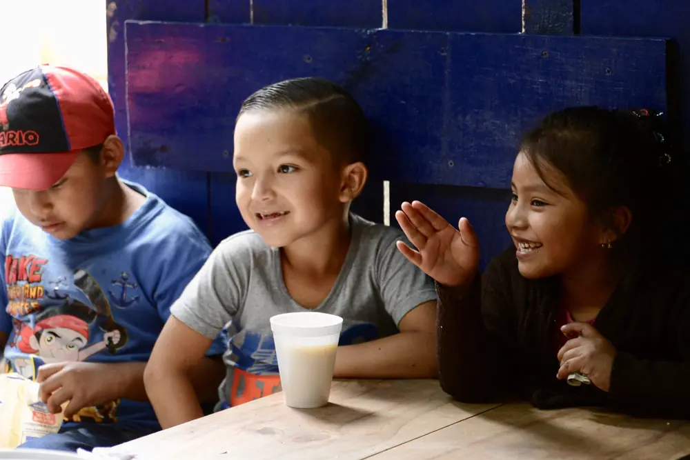 Three children laughing around a cup at a table