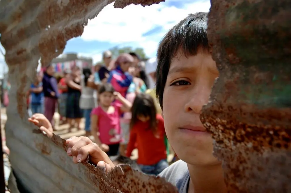 A boy's face framed by a hole in a rusted wall, a crowd beyond